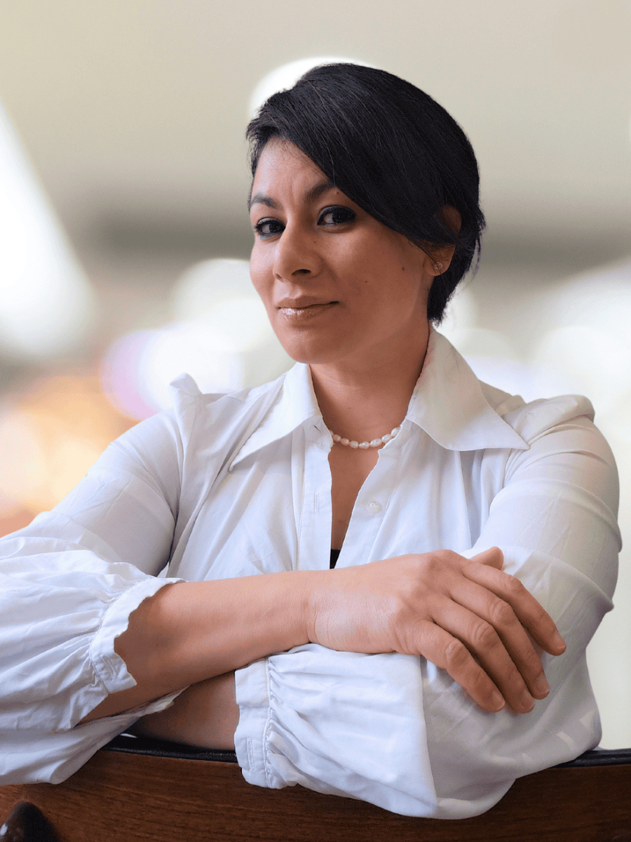 Ms. Nada Al-Hadithy, a woman with short dark hair, is sat with her arms leaning over her chair, wearing a white blouse with her arms crossed and a pearl necklace